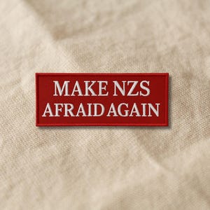 Könnte beinhalten: Ein rechteckiger, roter Aufnäher mit weiß gesticktem Text: "MAKE NZS AFRAID AGAIN". Der Aufnäher ist vor einem neutralen, strukturierten Hintergrund platziert.