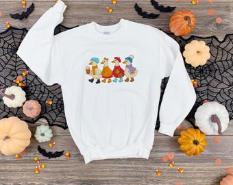 Puede incluir: Una sudadera blanca de manga larga con un estampado gráfico de cuatro patos vestidos con ropa de otoño. Los patos llevan gorros, bufandas y botas en tonos azul, naranja, rojo y morado, y uno de los patos sostiene una taza de café.