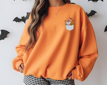 Puede incluir: Una sudadera naranja de cuello redondo con un pequeño gráfico de bolsillo blanco en el pecho izquierdo. El gráfico muestra un fantasma con un sombrero de bruja morado, una calabaza y pequeños murciélagos. La sudadera tiene mangas largas.