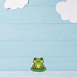 Puede incluir: Una pegatina de rana verde con una cara sonriente, sentada en una hoja de nenúfar. La pegatina está sobre un fondo de madera azul claro con dos nubes blancas.