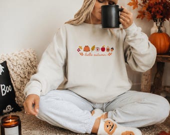 Puede incluir: Una sudadera beige de cuello redondo con un estampado gráfico de iconos de temática otoñal que incluye bayas, un suéter, una hoja, una taza, una manzana, una vela y una bellota, con el texto "hello autumn" escrito en cursiva en el pecho.