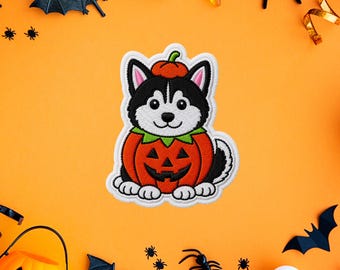 Puede incluir: Un parche bordado que muestra un cachorro de husky blanco y negro sentado dentro de una calabaza naranja tallada. El cachorro lleva un pequeño gorro de calabaza naranja en la cabeza con un detalle de cuello verde.