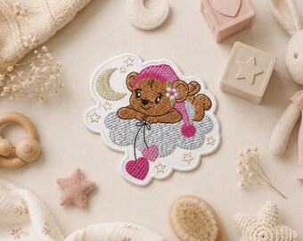Puede incluir: Un parche bordado que muestra un oso de peluche marrón con un gorro de dormir rosa con una pequeña flor, descansando sobre una nube gris. El oso sostiene dos globos rosas en forma de corazón. Una luna creciente dorada y estrellas rodean al oso.