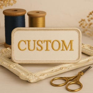 Puede incluir: Un parche rectangular crema bordado con la palabra "CUSTOM" en hilo dorado. El parche tiene un borde dorado. También se ven dos carretes de hilo y unas tijeras doradas.