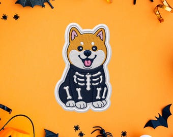 Puede incluir: Un parche bordado que muestra un perro Shiba Inu de color canela y blanco con un disfraz negro con un estampado de esqueleto blanco. El perro tiene orejas puntiagudas, ojos oscuros y una lengua rosa visible en su boca abierta, con borde blanco.