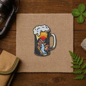 May include: A beer mug patch with a mountain and sunset design. The patch features a black mug with a handle, filled with a landscape scene of a sunset, mountains, and a waterfall. The top of the mug has white foam.