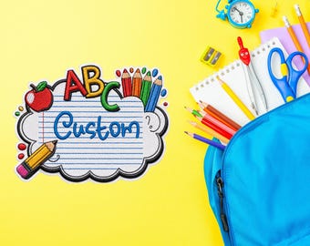 Puede incluir: Un parche bordado con forma de nube y papel rayado, que incluye el texto ABC y Custom, una manzana roja y un lápiz amarillo. A su lado hay una mochila azul, un cuaderno de espiral, un despertador azul, tijeras y lápices de colores.