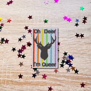 May include: A white rectangular patch with rainbow stripes and a black silhouette of a deer with antlers. The text "Oh Deer I'm Queer" is embroidered on the patch.
