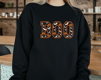 Puede incluir: Una sudadera negra de manga larga con la palabra BOO en letras grandes. Las letras tienen un borde naranja y están rellenas con un patrón blanco de pequeñas calabazas, murciélagos, estrellas y lunas crecientes sobre el fondo negro.