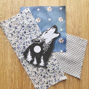 May include: A black and white wolf howling at the moon with a forest silhouette behind it. The wolf is on top of a white fabric with a blue and white floral pattern. There is a blue fabric with white polka dots and a white fabric with blue polka dots behind the wolf.