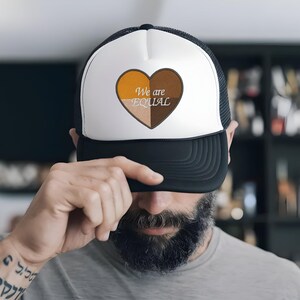 Puede incluir: Gorra de camionero en blanco y negro con un panel frontal blanco que presenta un diseño de corazón dividido en cuatro secciones marrones. Las palabras "We are EQUAL" están bordadas en blanco dentro del corazón.