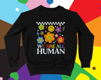 Puede incluir: Sudadera negra con un diseño floral y de caritas sonrientes. El texto "WE ARE ALL HUMAN" está en blanco. El diseño presenta flores, rayos y un patrón de tablero de ajedrez.