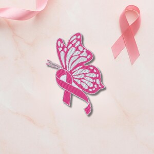 Könnte beinhalten: Ein gestickter rosa und weißer Schmetterlingsaufnäher mit einem rosa Band, das das Bewusstsein für Brustkrebs symbolisiert. Der Schmetterling hat detaillierte Flügelmuster. Ein rosa Band und ein rosa Band sind ebenfalls auf einem hellrosa Marmorhintergrund vorhanden.