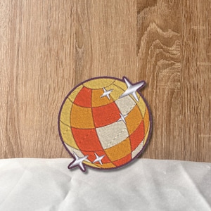 May include: A red, orange and white disco ball with silver glitter stars. The disco ball is embroidered on a fabric patch.