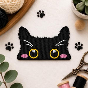 Può includere: Una toppa ricamata a forma di muso di gatto nero con grandi occhi gialli con riflessi bianchi, guance rosa e baffi bianchi. La toppa ha orecchie a punta ed è circondata da piccole impronte di zampe ricamate in nero.