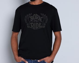 Puede incluir: Camiseta negra con el texto "BORN TO RIDE" en una fuente estilizada con acentos en forma de alas. La camiseta de manga corta está hecha de un material suave. El modelo lleva vaqueros azules.