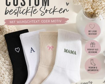 Puede incluir: Cuatro calcetines acanalados en negro, blanco, beige y blanco. El calcetín negro tiene un corazón blanco, el blanco una letra A azul, el beige un lazo rosa y el blanco el texto bordado en verde MAMA.