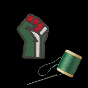 Könnte beinhalten: Ein gestickter Aufnäher mit einer erhobenen Faust in Grün, Rot und Weiß, umrandet in Grau. Eine Spule grünen Fadens und eine Nadel sind auf schwarzem Hintergrund daneben platziert. Das Patch-Design ist ein Symbol der Solidarität.