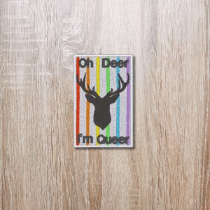 May include: A white patch with a rainbow stripe pattern and a black silhouette of a deer with antlers. The text "Oh Deer I'm Queer" is written in black.