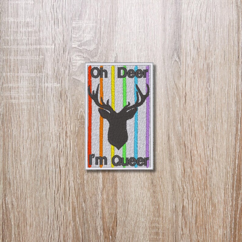May include: A white patch with a rainbow stripe pattern and a black silhouette of a deer with antlers. The text "Oh Deer I'm Queer" is written in black.