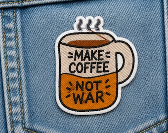 Puede incluir: Un parche bordado con forma de taza de café con borde blanco, que presenta una mitad superior beige y una mitad inferior naranja. El texto "MAKE COFFEE" está en negro en la sección beige, y "NOT WAR" está en negro en la sección naranja.
