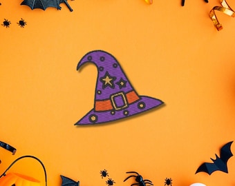 Puede incluir: Un parche de sombrero de bruja morado con una estrella dorada, una hebilla dorada sobre una banda naranja y pequeños círculos dorados. El sombrero tiene una punta curva y un ala ancha, con una textura que imita el bordado sobre un fondo naranja.