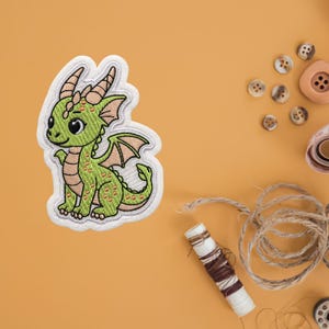 May include: Embroidered patch featuring a friendly green dragon with a white border. The dragon has large eyes, small wings, and a detailed design. The patch is likely for clothing or accessories.