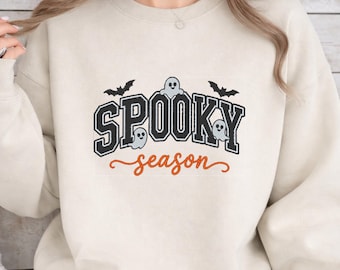 Puede incluir: Una sudadera de cuello redondo color crema con un diseño gráfico que presenta el texto SPOOKY en letras negras de estilo universitario y season en cursiva naranja. El diseño incluye tres pequeños fantasmas azul claro y dos murciélagos negros.