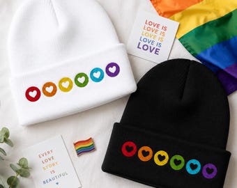 Puede incluir: Dos gorros, uno blanco y otro negro, cada uno bordado con una fila de corazones de colores del arcoíris. También se ven un pequeño pin con una bandera arcoíris y tarjetas con el texto "LOVE IS LOVE" y "EVERY LOVE STORY IS BEAUTIFUL".