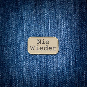May include: A rectangular patch with the German words "Nie Wieder" in black text, stitched onto a textured, light tan background. The patch is set against a backdrop of blue denim fabric.