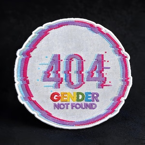 Pode incluir: Um patch redondo branco com um design de efeito glitch. O texto "404 GENDER NOT FOUND" é exibido em letras coloridas e pixeladas. O patch tem uma superfície texturizada e uma borda branca com detalhes em rosa e azul.