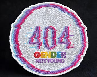 Puede incluir: Un parche redondo blanco con un diseño de efecto glitch. El texto "404 GENDER NOT FOUND" se muestra en letras coloridas y pixeladas. El parche tiene una superficie texturizada y un borde blanco con acentos rosas y azules.