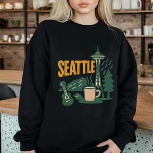 Pode incluir: Camisola preta com um design de Seattle. O design inclui a palavra "SEATTLE" em amarelo, uma ilustração da Space Needle, uma guitarra, montanhas, uma chávena de café e árvores.