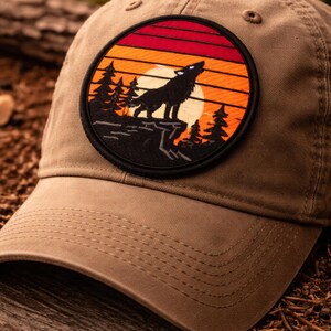 May include: A tan baseball cap with a circular patch showing a sunset scene. The patch features a black wolf howling at a yellow sun, with pine trees and a red, orange, and yellow sky. The cap has a curved peak and a fabric strap.