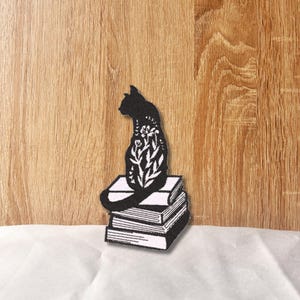 Könnte beinhalten: Schwarze Silhouette einer Katze mit floralen Details, die auf einem Stapel Bücher sitzt.