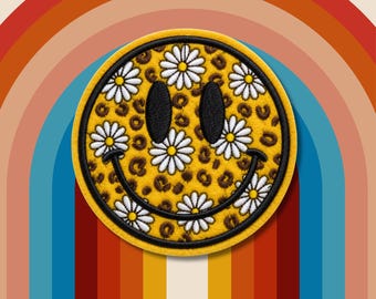 Puede incluir: Un parche bordado circular que presenta una cara sonriente amarilla con un patrón de estampado de leopardo marrón. La cara está decorada con varias flores de margarita blancas con centros amarillos, dos ojos ovalados negros y una boca negra curva.
