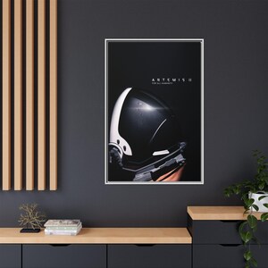 May include: A framed poster featuring a close-up of a black and white astronaut helmet. The text "ARTEMIS II FOR ALL HUMANITY" is displayed at the top. The poster is mounted on a dark gray wall above a wooden cabinet.
