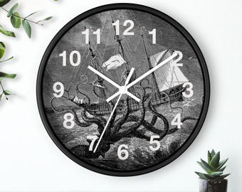 May include: A round wall clock with a black frame and white hands. The clock face features a black and white illustration of a large sailing ship being attacked by a giant octopus in the ocean. Large white numbers mark the hours on the face.