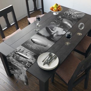 May include: Black and white table runner with a vintage airship design. The runner is placed on a dark wood table with white plates and silverware.
