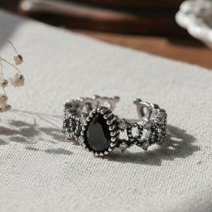 May include: A silver ring with an adjustable band. It has a teardrop-shaped black stone at the centre, encircled by small, clear stones and decorative filigree. The ring is set against a neutral background.
