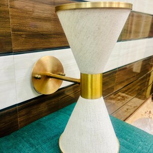 Könnte beinhalten: Eine goldene Wandleuchte mit einem weißen Stoffschirm. Der Schirm ist wie eine Sanduhr geformt, mit einem breiteren oberen und unteren Teil und einer schmaleren Mitte. Die Leuchte hat ein schlichtes, modernes Design.