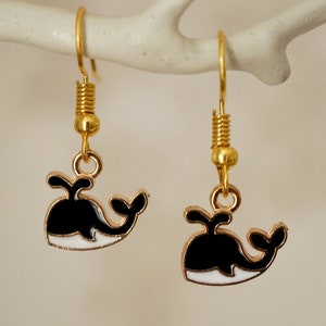 May include: A pair of gold dangle earrings with black and white whale charms. The whales have a white belly and a black body with a white wave underneath.