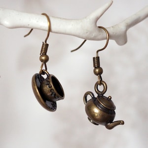 May include: A pair of antique bronze-colored earrings. One earring features a teacup and saucer, the other a teapot. Both earrings hang from a simple hook and coil design. The earrings are suspended against a white background.