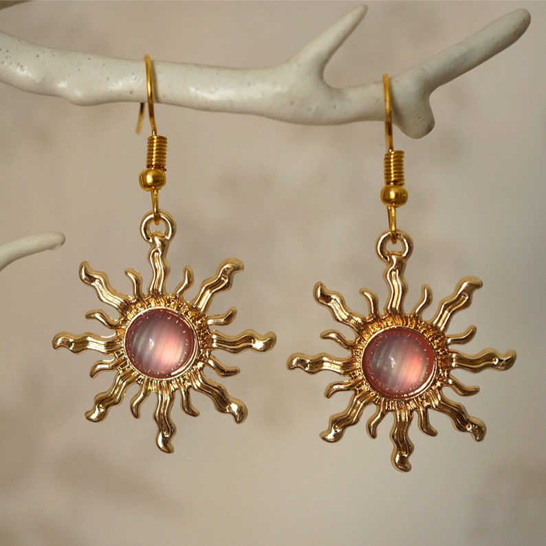 K&ouml;nnte beinhalten: Goldene Sonnenohrringe mit rosafarbenen, schillernden Glaszentren. Die Ohrringe haben ein zartes, feminines Design.