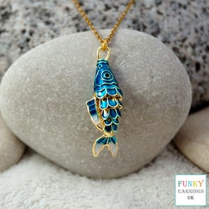 May include: A gold-toned necklace featuring a pendant shaped like a fish with vibrant blue enamel scales and gold trim. The fish pendant hangs from a delicate gold chain. A small square logo in the corner displays the text FUNKY EARRINGS UK.