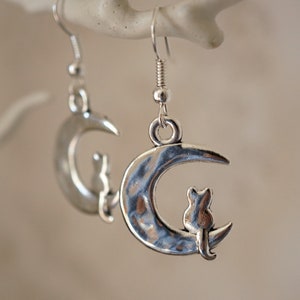 May include: Silver earrings with a crescent moon design and a small cat silhouette sitting on the moon.