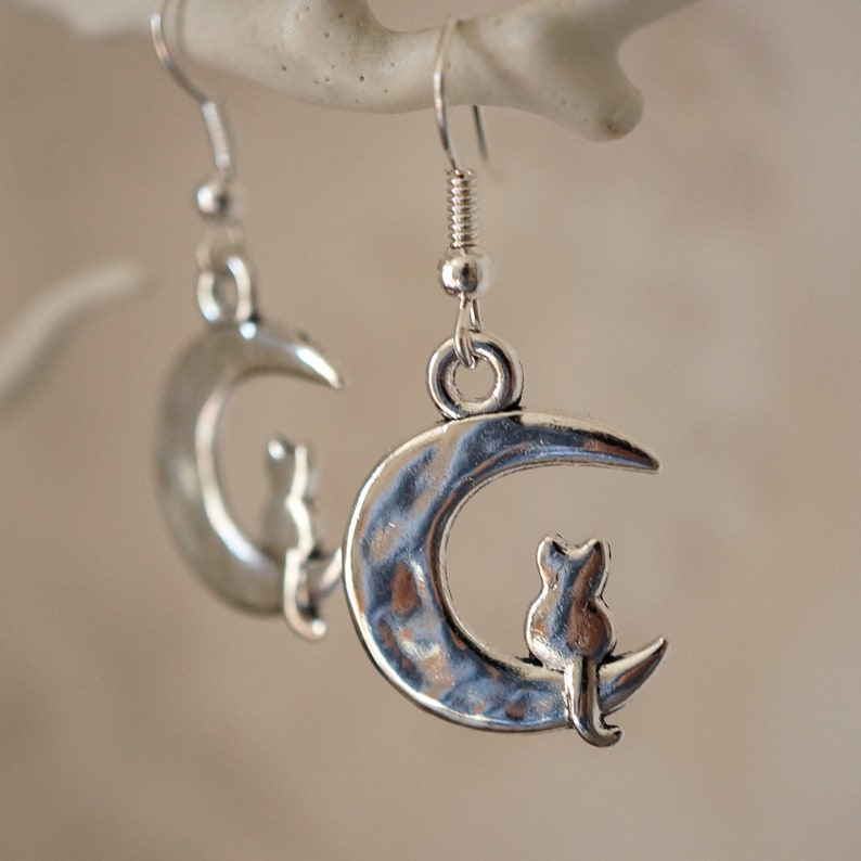 May include: Silver earrings with a crescent moon design and a small cat silhouette sitting on the moon.