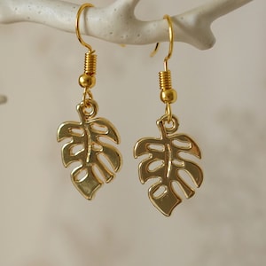 May include: A pair of gold-tone earrings featuring a cutout monstera leaf design. The earrings are dangle style with a hook fastening.