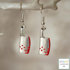 May include: A pair of silver earrings shaped like a bloody butcher knife. The earrings have red paint on the blade and red dots on the handle.