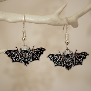 May include: A pair of silver dangle earrings featuring a black bat design with celestial details. The bat has a starry night sky pattern with moons and stars.